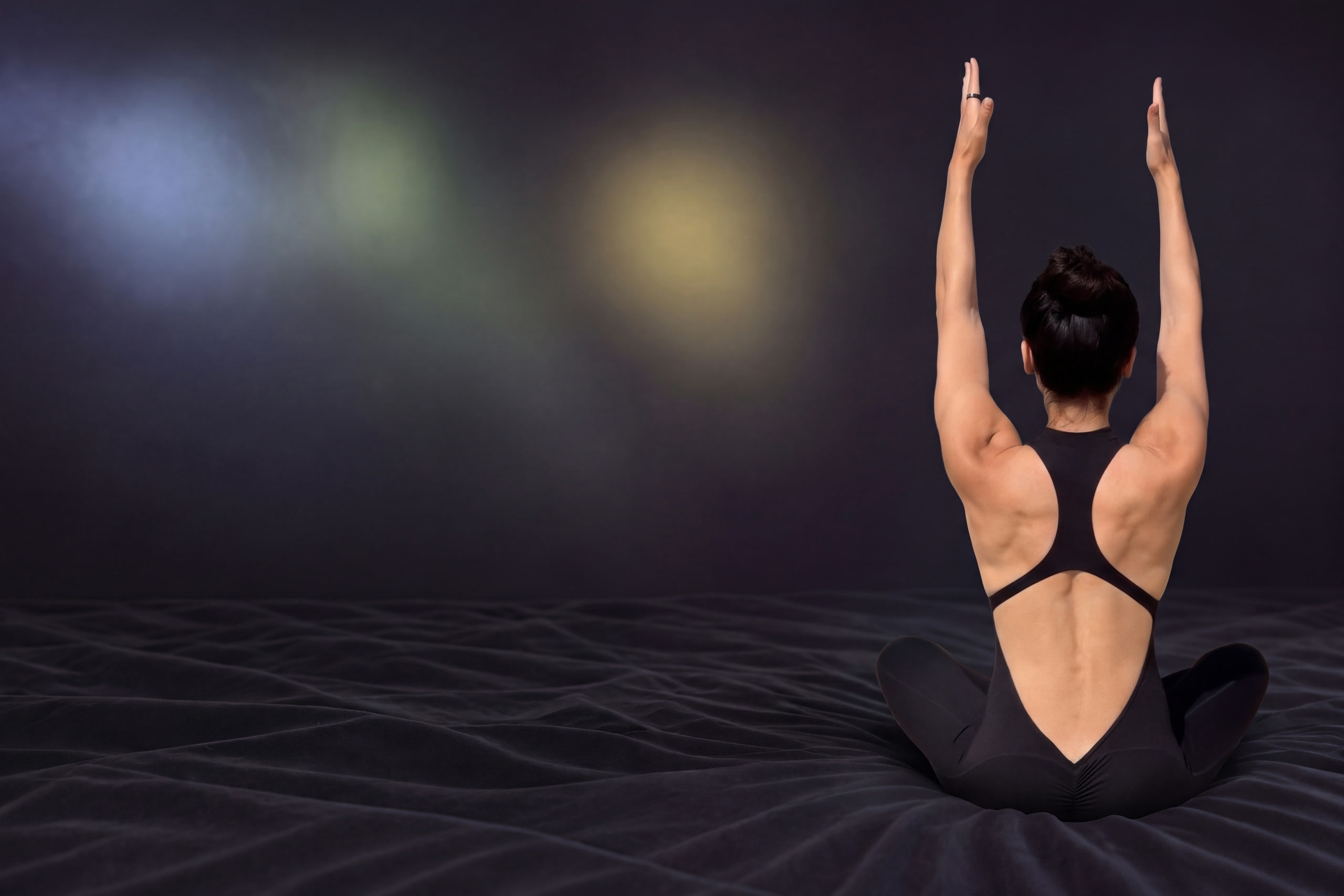 Karina from behind, sitting cross-legged on dark sheets, arms raised, in a black open-back jumpsuit